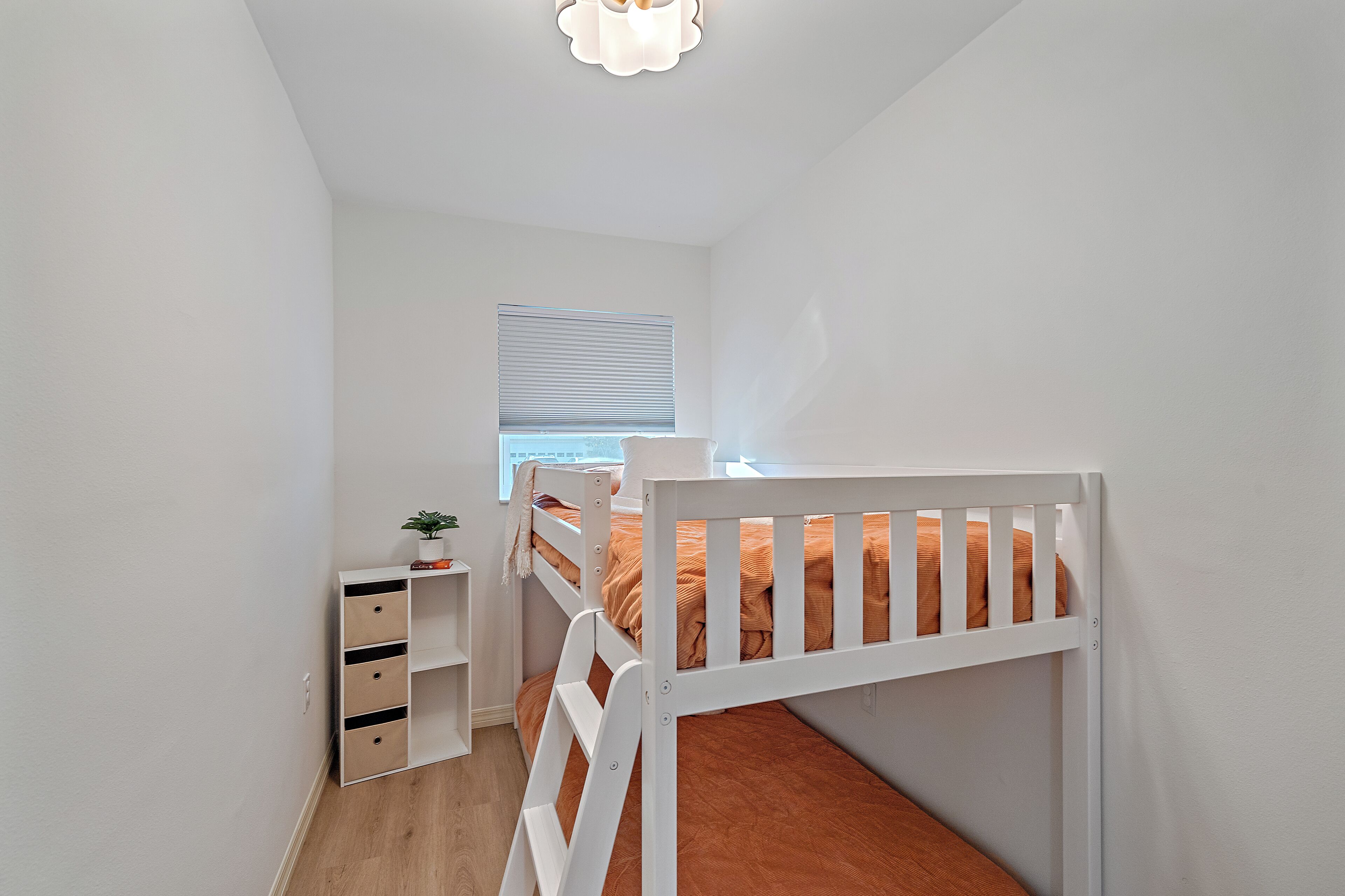 A bunk bed with a white frame and orange bedding, a small bedside table with a plant, and a window with blinds.
