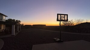 Een buitenbasketbalveld met een basket, omringd door een verhard gebied en een gebouw aan één kant.