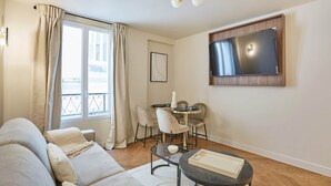 A living room with a sofa, a coffee table, a TV mounted on the wall, and a dining area with chairs and a table. - New Apartment Center of Paris/le Marais (Paris)