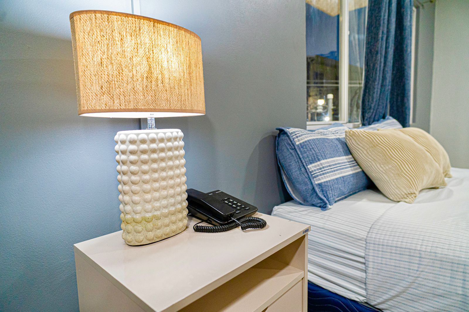 A hotel room with a bedside lamp, a phone, and a bed with pillows.