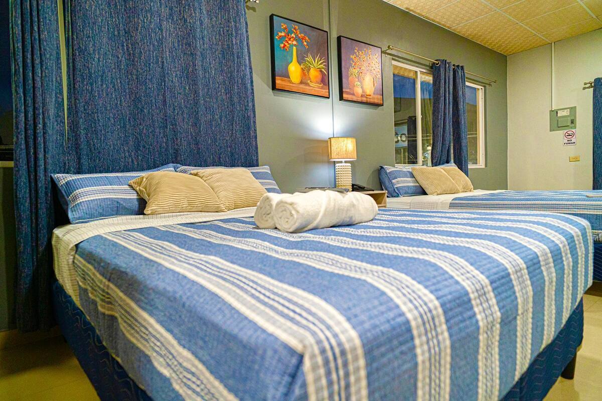 A hotel room with two beds, blue and white striped bedding, a lamp, and two framed floral paintings on the wall.