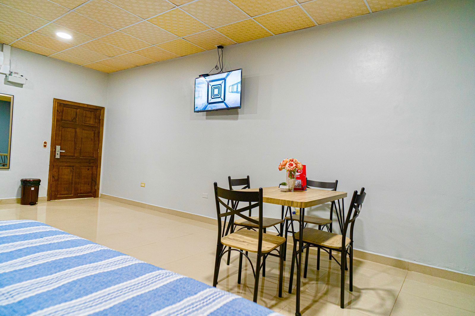 A hotel room with a bed, a dining table with chairs, a TV mounted on the wall, and a trash bin.