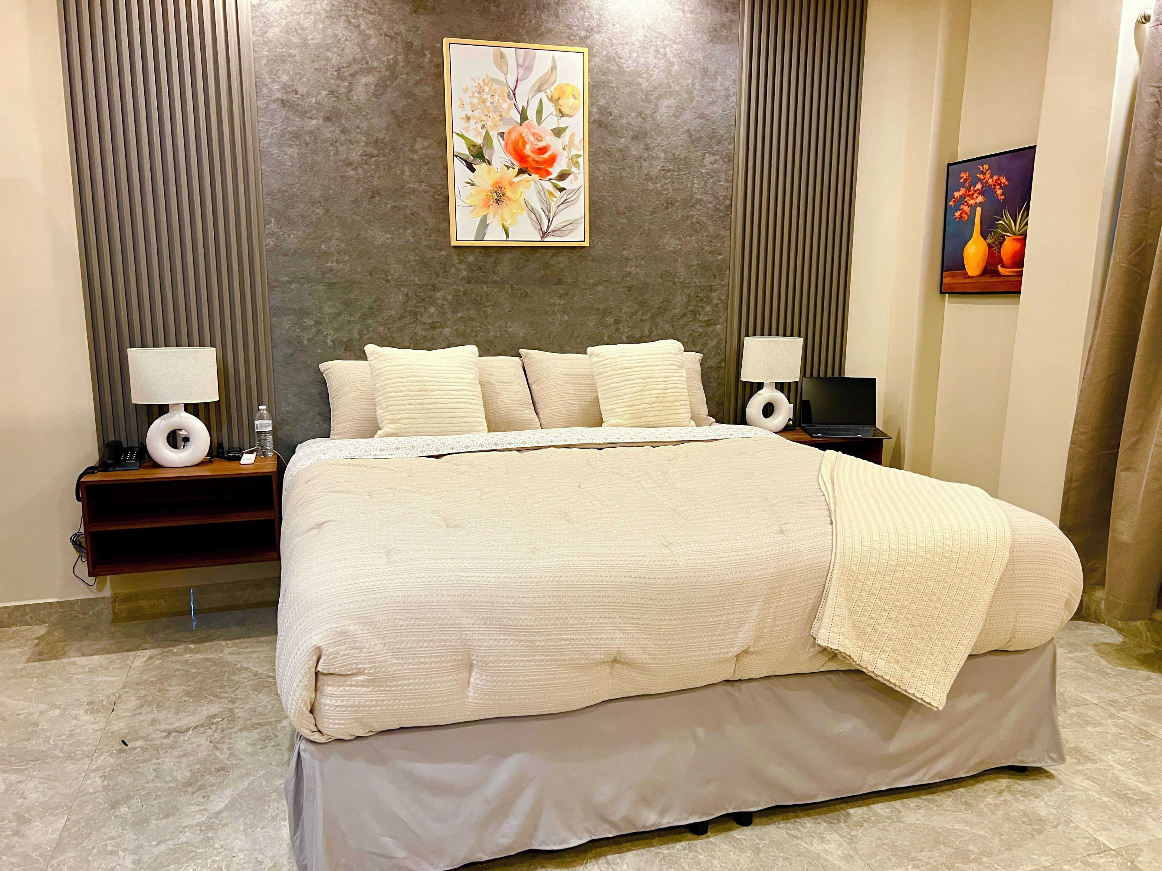 A neatly made bed with a white comforter and pillows, a bedside table with a lamp, a wall-mounted painting, and a small table with a laptop.