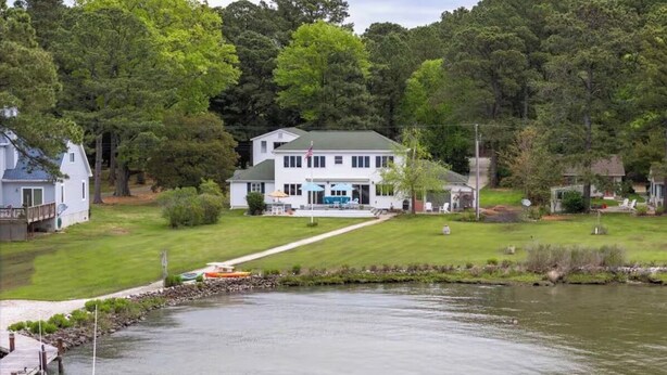 Una casa grande con césped verde y un muelle que se adentra en un lago, rodeada de árboles frondosos.