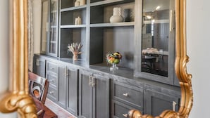 A dark wood cabinet with multiple shelves and drawers, a glass-top table with a fruit bowl, and a gold-framed mirror.