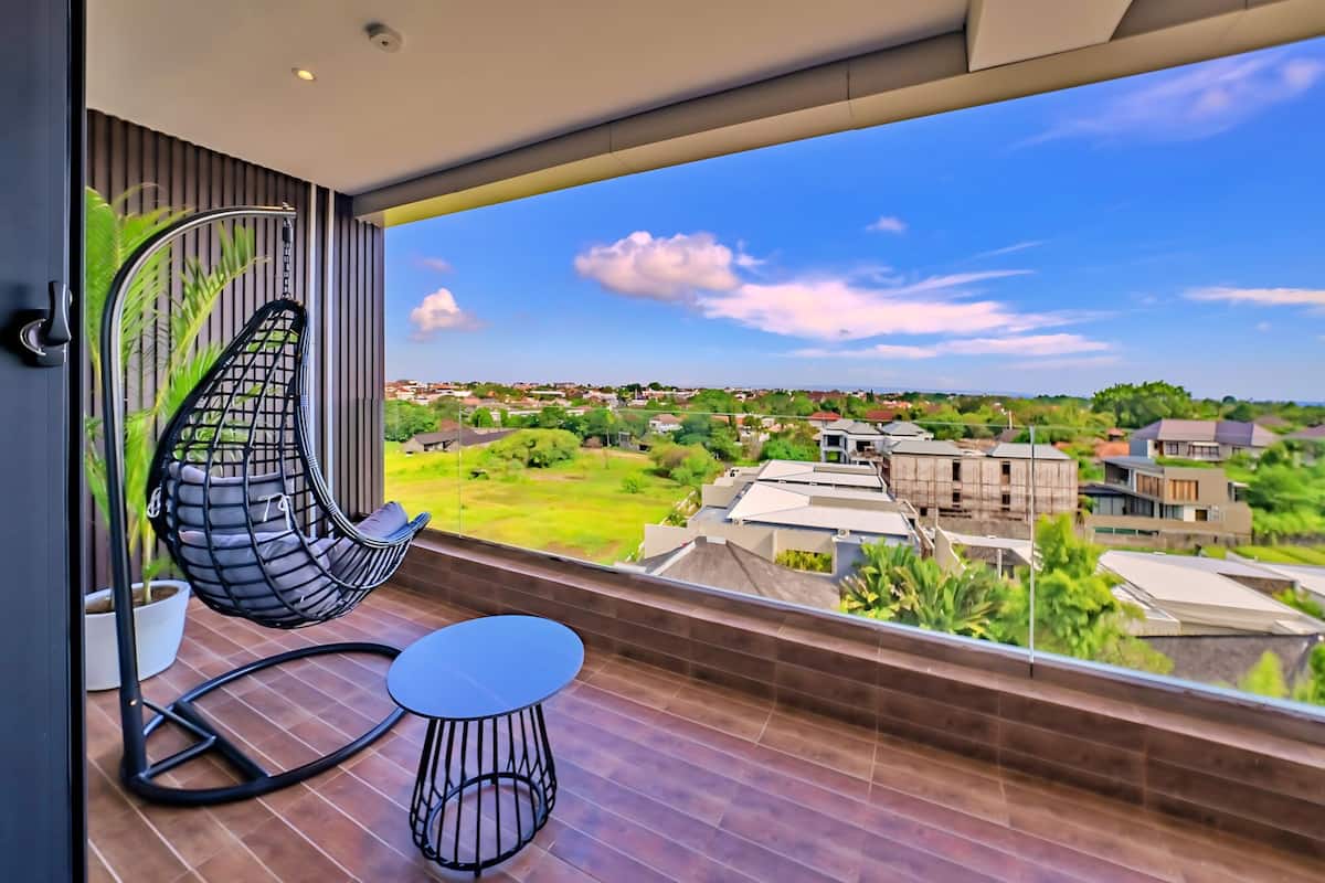 A balcony with a hanging chair and a small table, overlooking a residential area with houses and greenery.