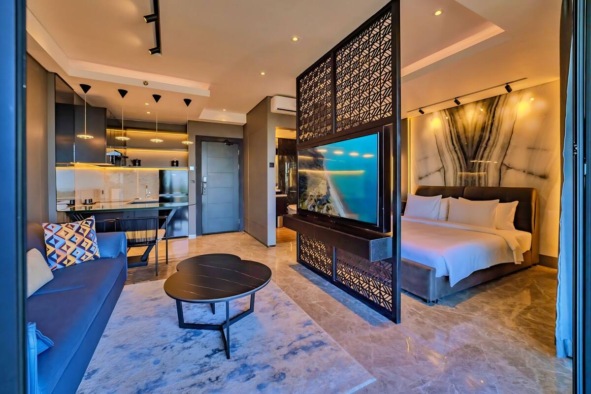 A modern hotel room with a large bed, a TV mounted on a wall, a dining area with a table and chairs, and a separate seating area with a sofa and a coffee table.