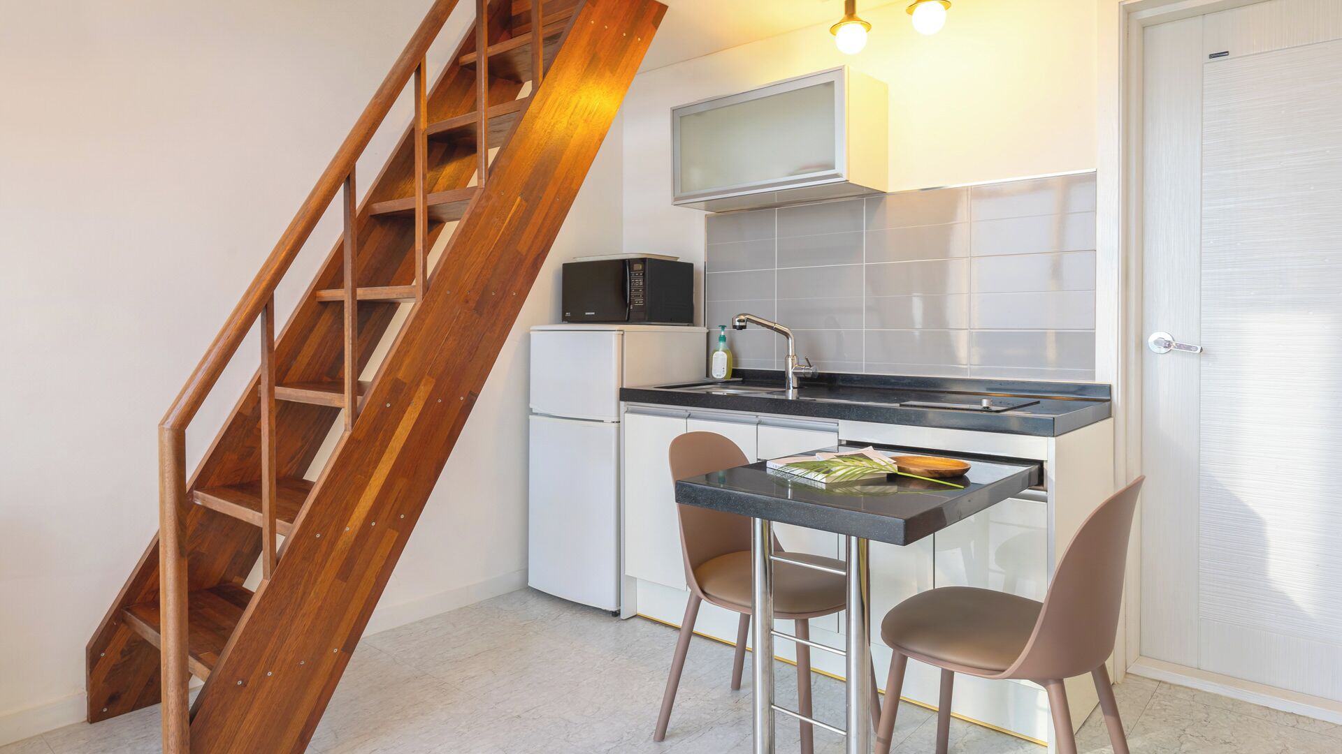 A compact kitchen with a wooden staircase, a microwave, a sink, and two chairs.