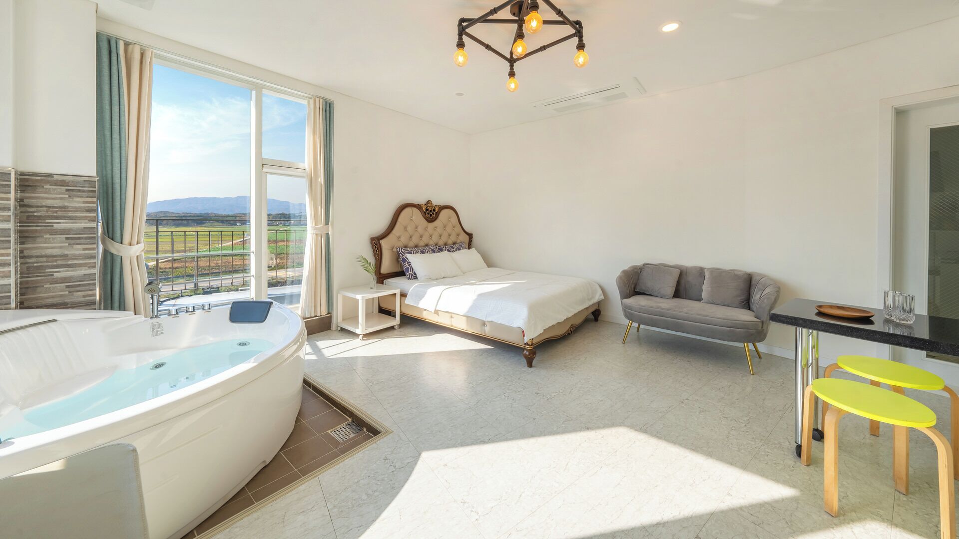 A modern hotel room with a large bed, a sitting area with a sofa and chairs, a small table, a bathtub, and a balcony with a view.