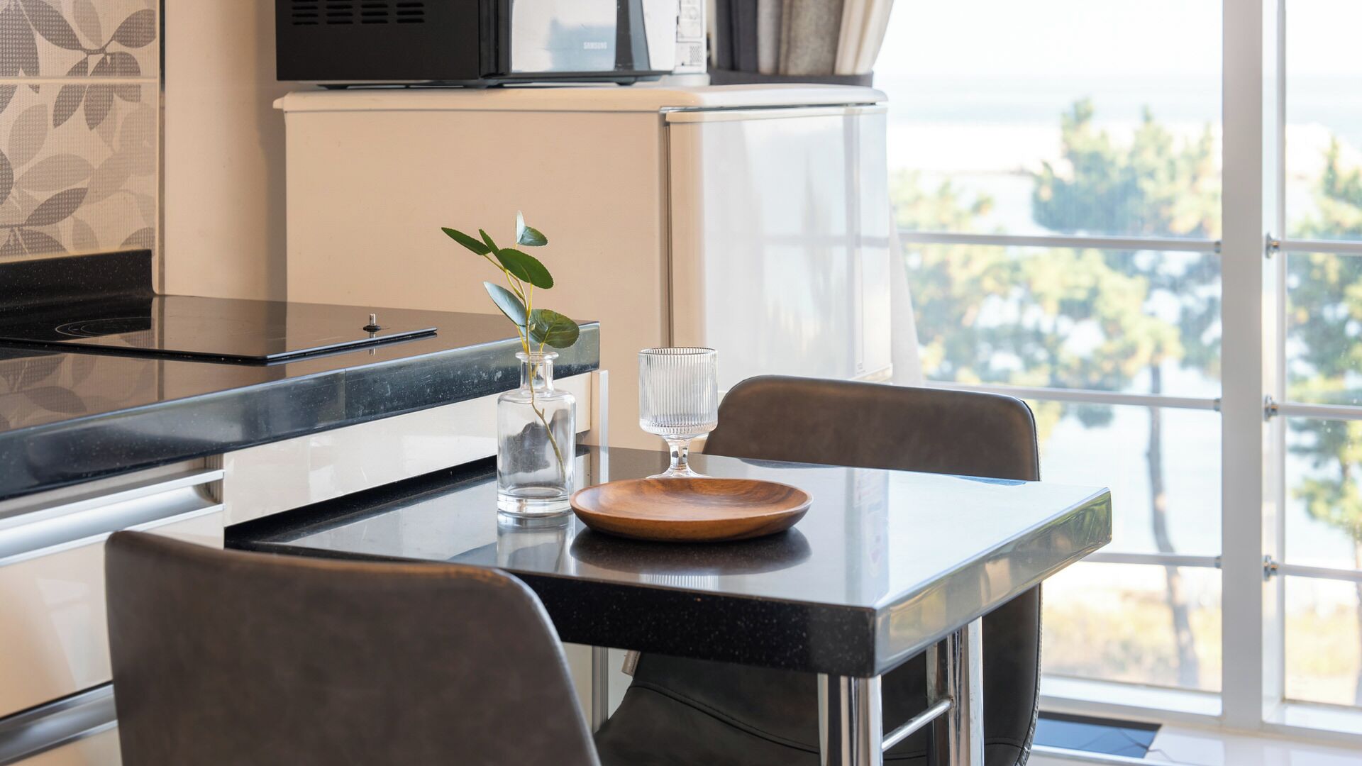 A modern kitchen with a dining table, chairs, a glass vase with a plant, and a view of the outdoors through large windows.