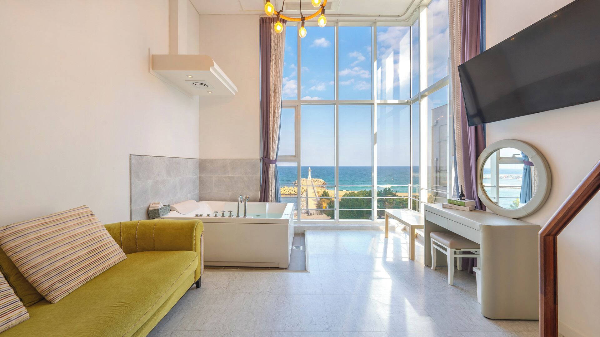A modern hotel room with a large window, a sofa, a bathtub, and a TV.
