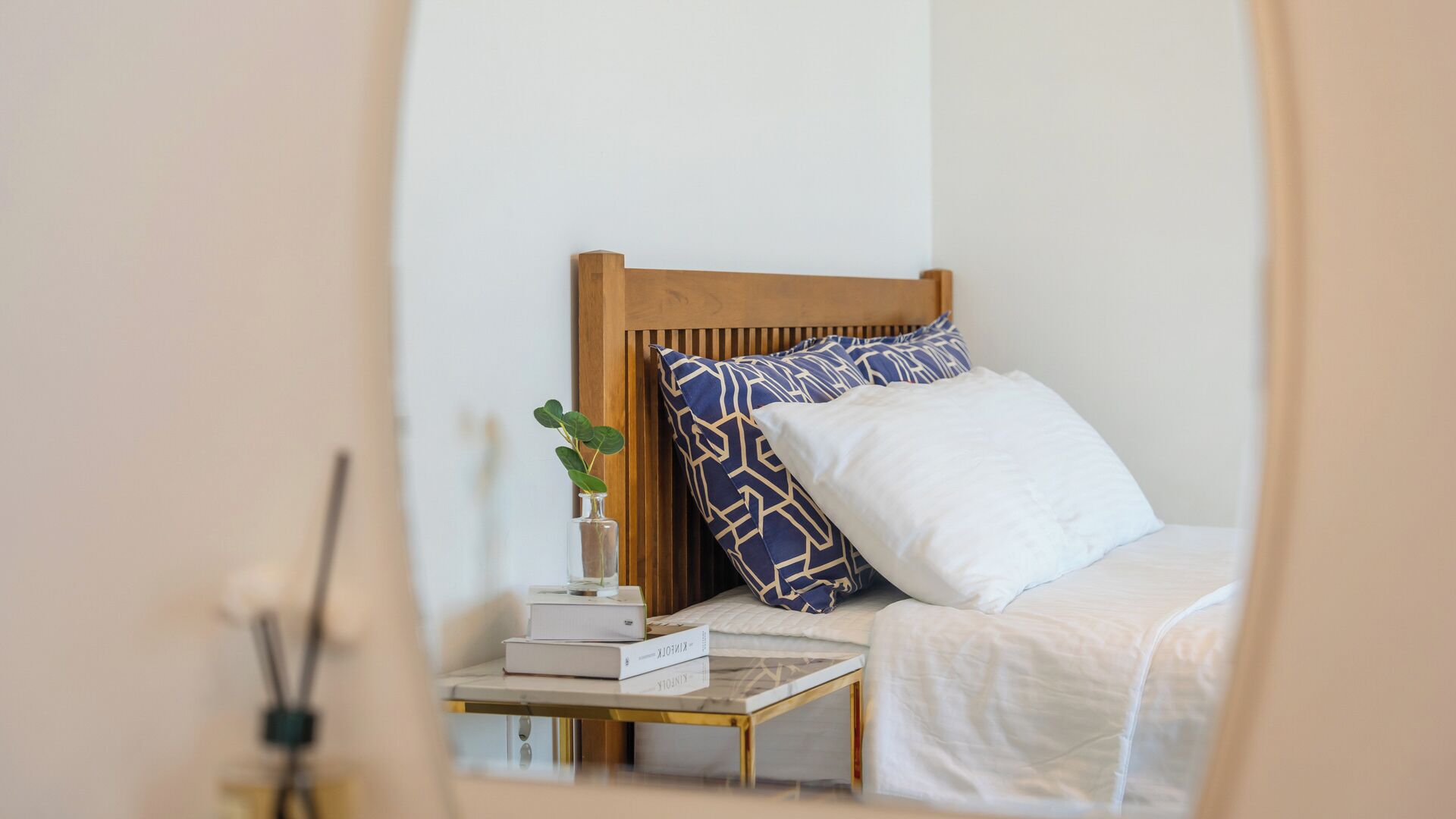 Un lit bien fait, avec une tête de lit en bois, une table de chevet sur laquelle sont posés des livres et une plante, et un oreiller blanc.