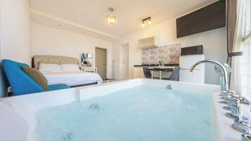 A hotel room with a large bathtub, a bed with blue and brown pillows, a desk with a computer, and a kitchen area.