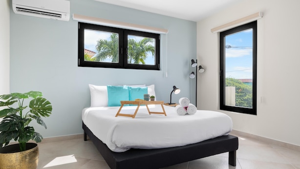 A modern bedroom with a bed, a potted plant, and a window with a view of palm trees. - Modern NEW 2BR1BA ~ Nature Escape ~ Great Location (Paradera)