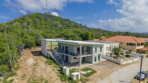 A modern two-story house with a balcony, surrounded by a lush green landscape and a hill. - Modern NEW 2BR1BA ~ Nature Escape ~ Great Location (Paradera)