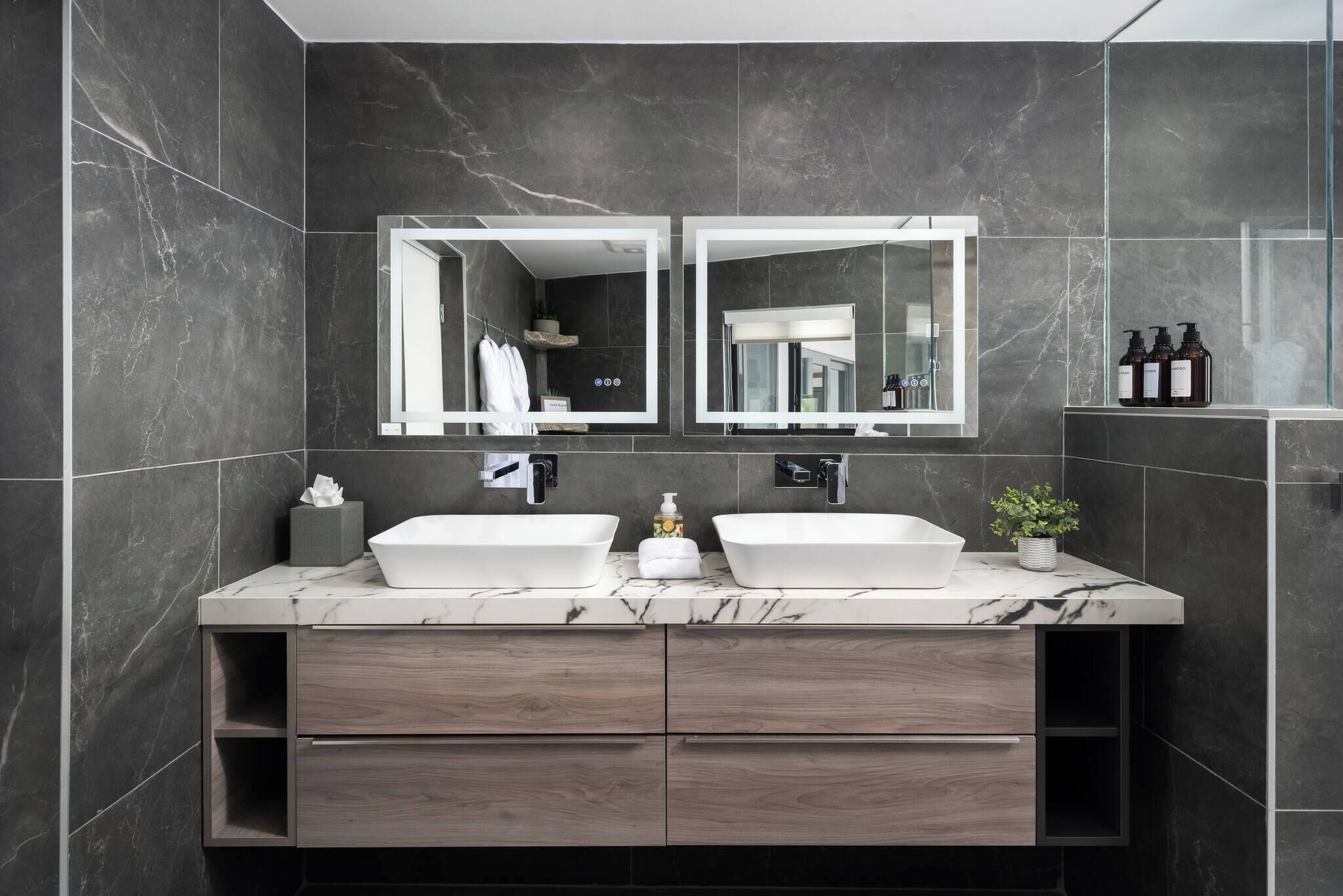 Une salle de bain moderne avec deux vasques, un grand miroir et des carreaux de marbre foncé.
