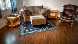 A living room with a brown leather armchair, a brown sofa, a small wooden side table, a glass coffee table, and a patterned rug.