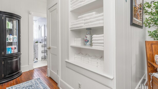 Un couloir avec une porte blanche donnant sur une salle de bain, un meuble noir avec des portes en verre et une étagère à serviettes blanche.