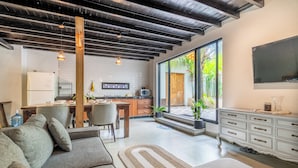 Een moderne woonkamer met een bank, een eethoek met een tafel en stoelen, een keuken met apparatuur, en een groot raam met uitzicht op groen.
