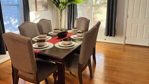 A dining room with a wooden table set for four, surrounded by four chairs. A vase with green plants is on the table. - Ocean View Beach House. Sleeps 8 Coastal Luxe. Seabreeze! (Norfolk)