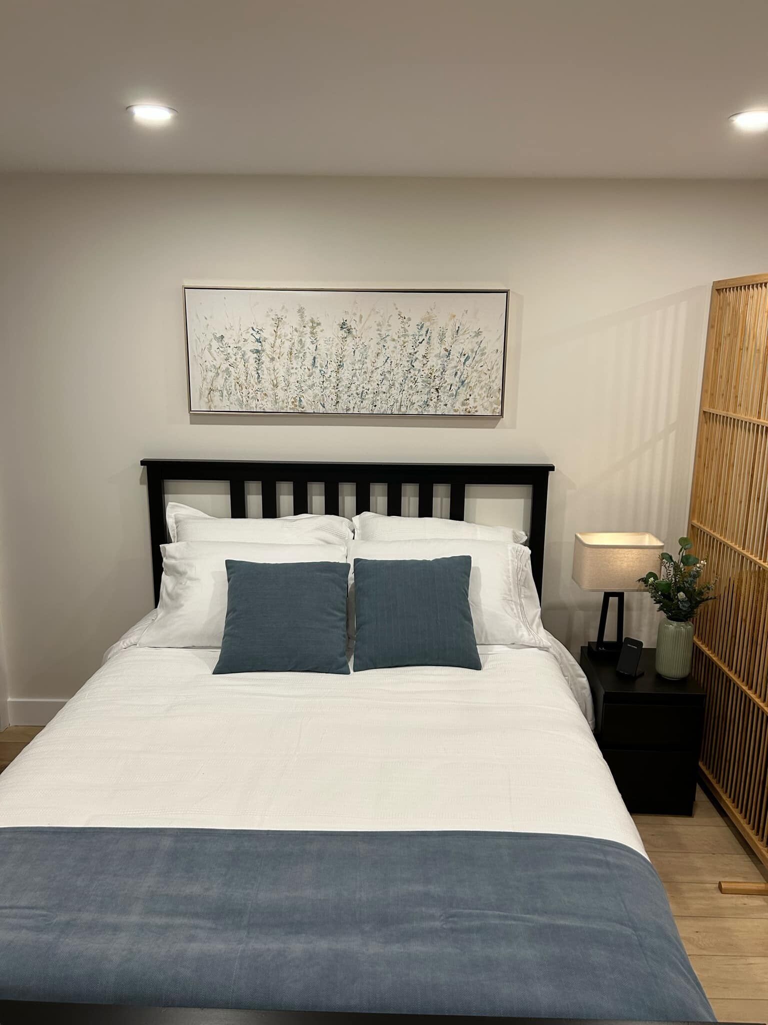 A neatly made bed with white and blue bedding, a wall-mounted artwork, and a bedside table with a lamp and a small plant.