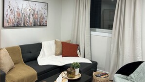Een moderne woonkamer met een zwarte bank, een klein bijzettafeltje, een witte plaid en een schilderij aan de muur.