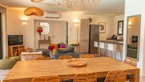 A modern living room with a wooden dining table, wicker chairs, a grey sofa, and a kitchen area with a refrigerator and bar stools.