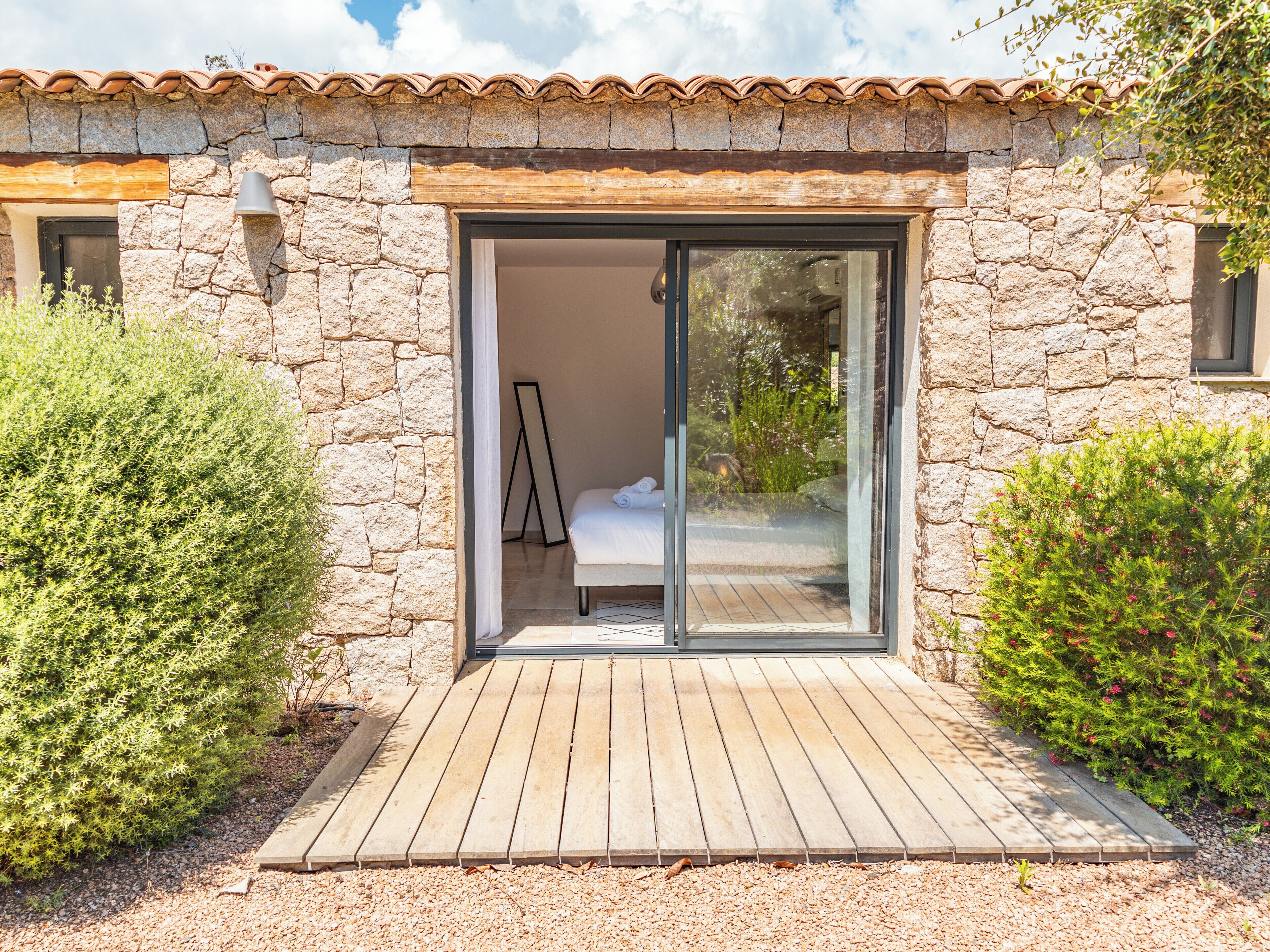 Un edificio de piedra con un porche de madera, un ventanal corredero que da a un dormitorio y un pequeño jardín.