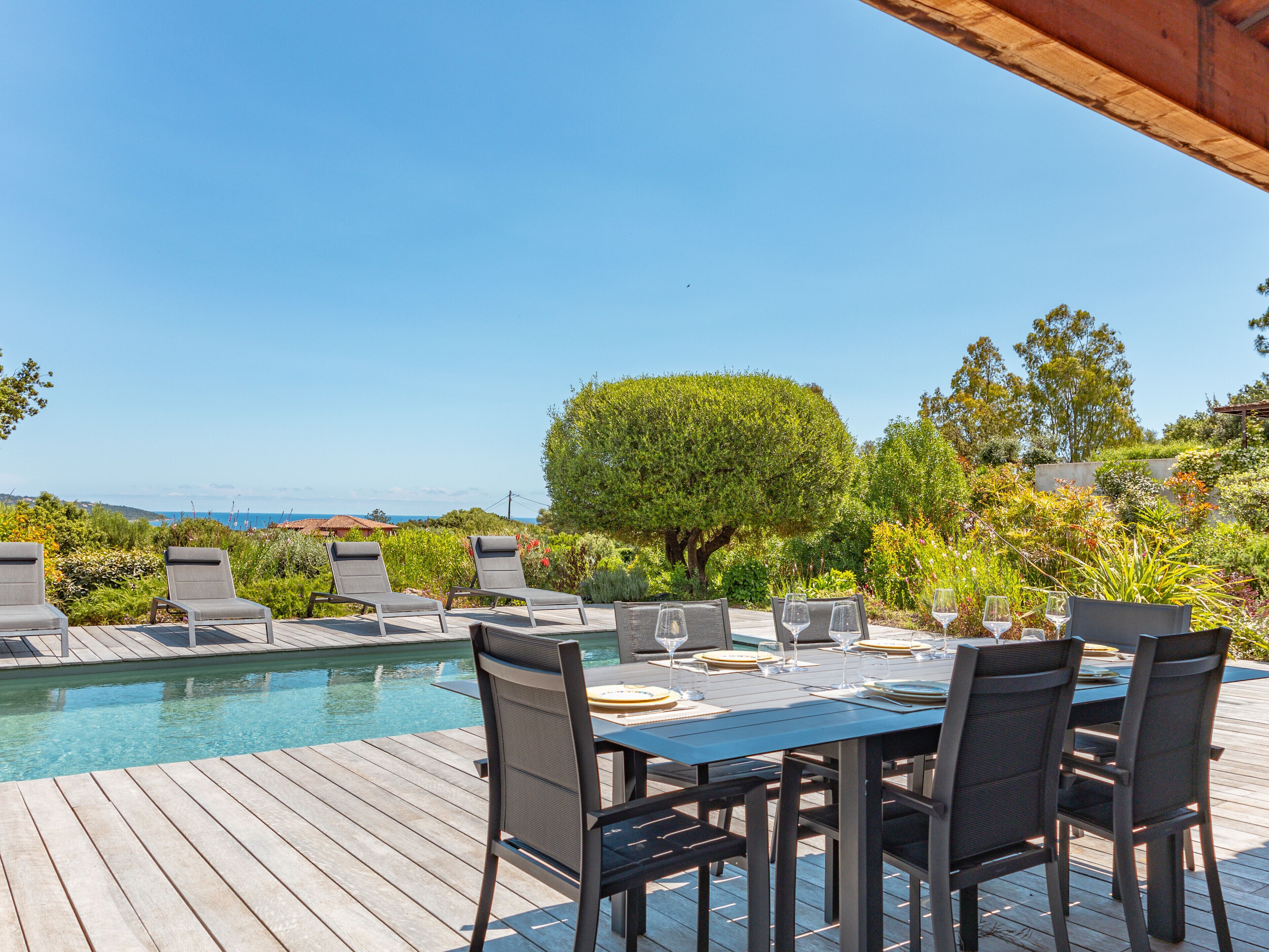 A poolside dining area with a view of the ocean, surrounded by lush greenery and a wooden deck.