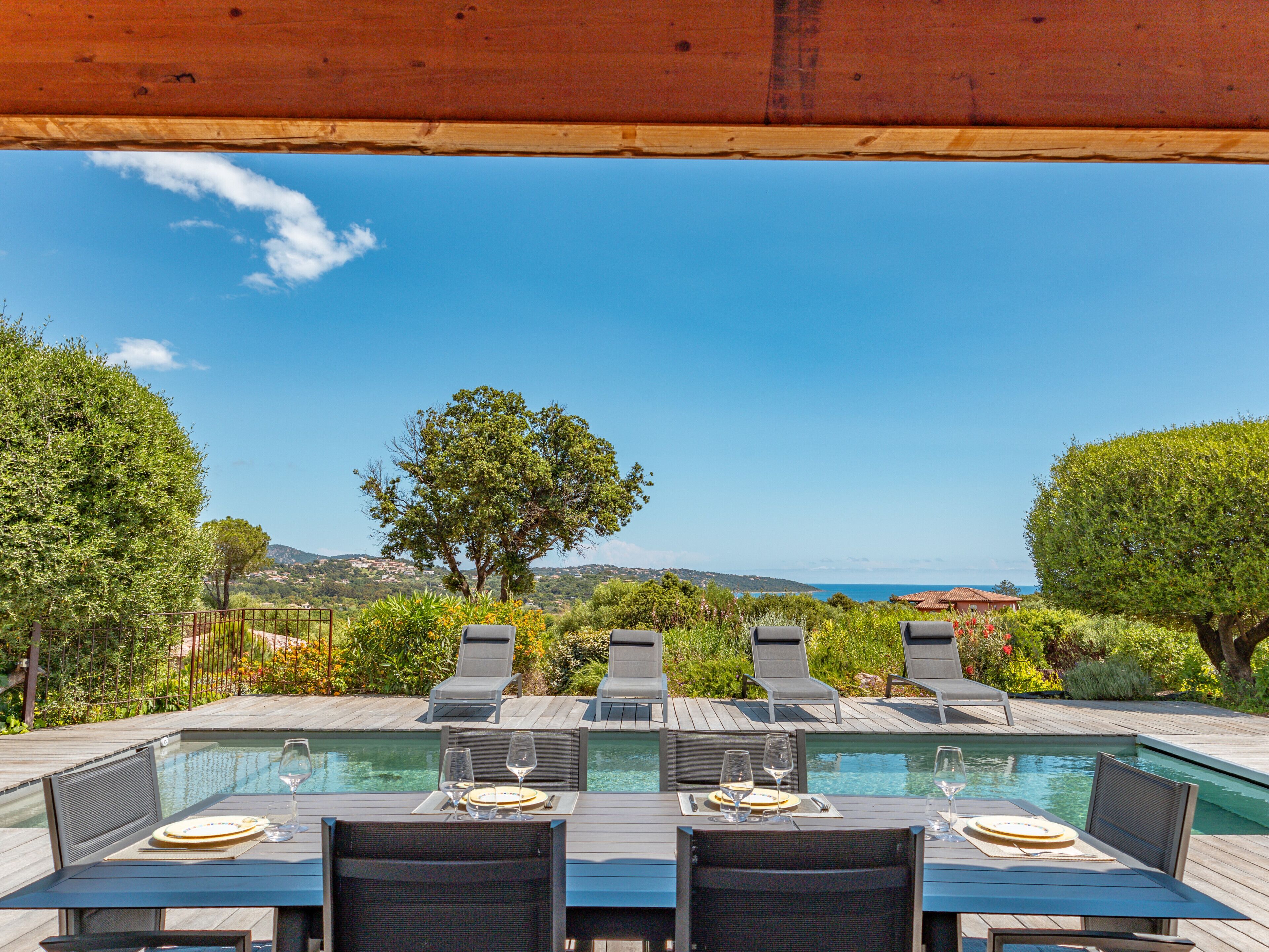 A poolside dining area with tables and chairs, overlooking a pool and a scenic view of hills and the sea.