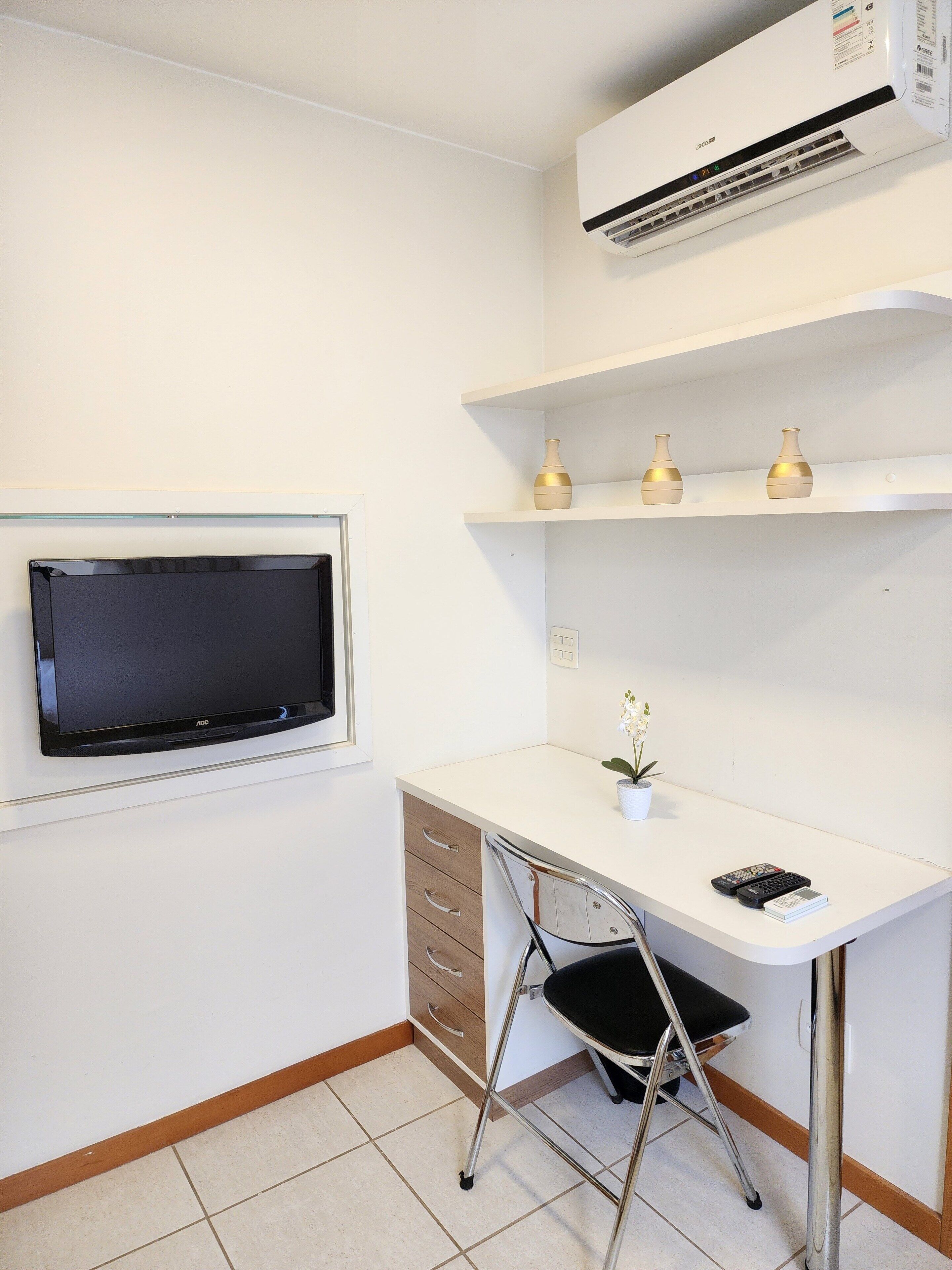 A compact office space with a white desk, a chair, a television, an air conditioner, and a small potted plant.