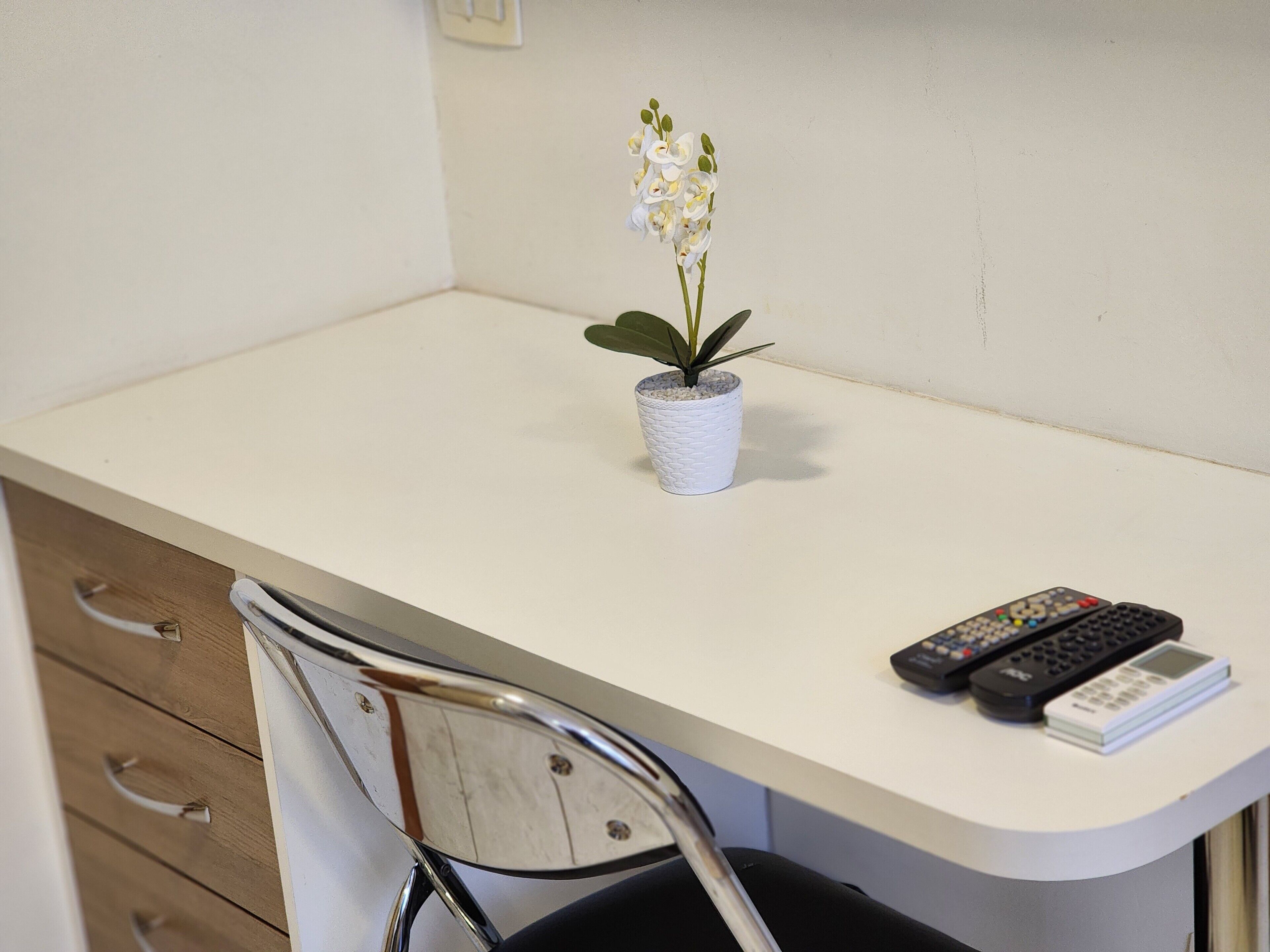 Een wit bureau met een bloeiende orchidee, een zwarte afstandsbediening en een wit apparaat.
