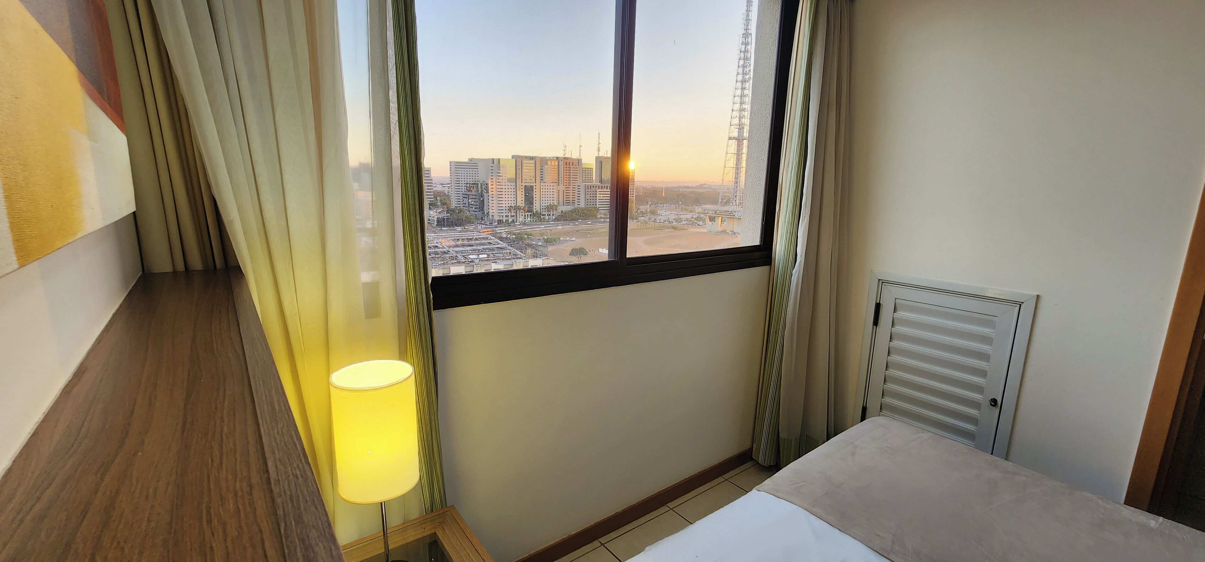 Una habitación de hotel con cama, mesita de noche, lámpara y ventana con vista a la ciudad.