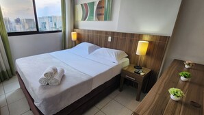 Een hotelkamer met een net opgemaakt bed, twee bedlampjes, een houten nachtkastje met een afstandsbediening, en een kleine tafel met potplanten.