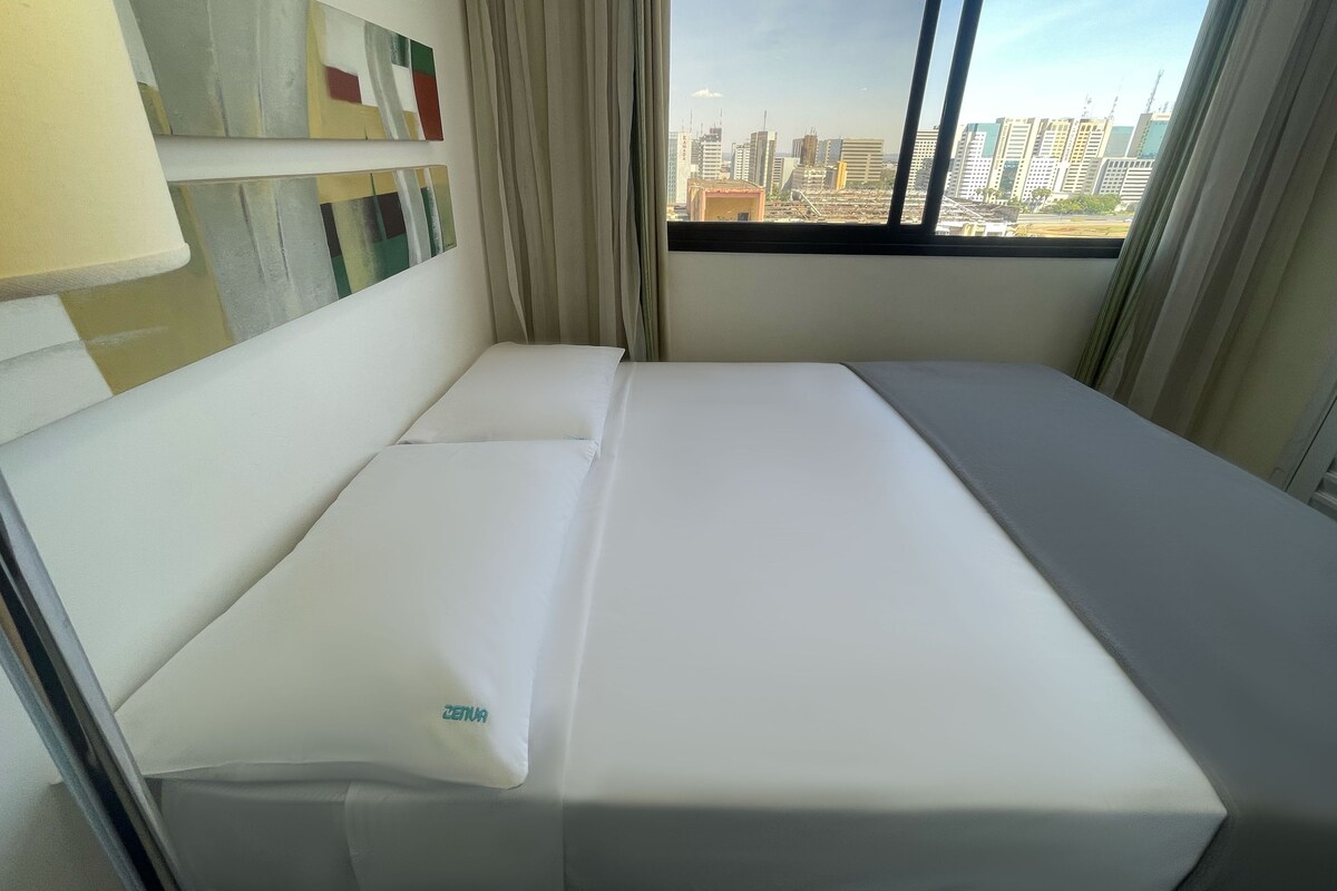 A hotel room with a bed, a window with a city view, and a painting on the wall.
