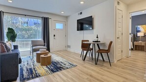 Ein modernes Wohnzimmer mit einer Couch, einem Couchtisch und Sesseln, einem Essbereich mit Tisch und Stühlen, einem an der Wand montierten Fernseher und einem großen Fenster mit Jalousien.