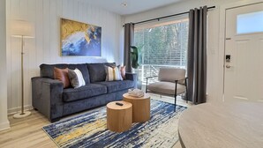 Ein modernes Wohnzimmer mit einem grauen Sofa, einem runden Couchtisch und einem großen Fenster mit Jalousien.