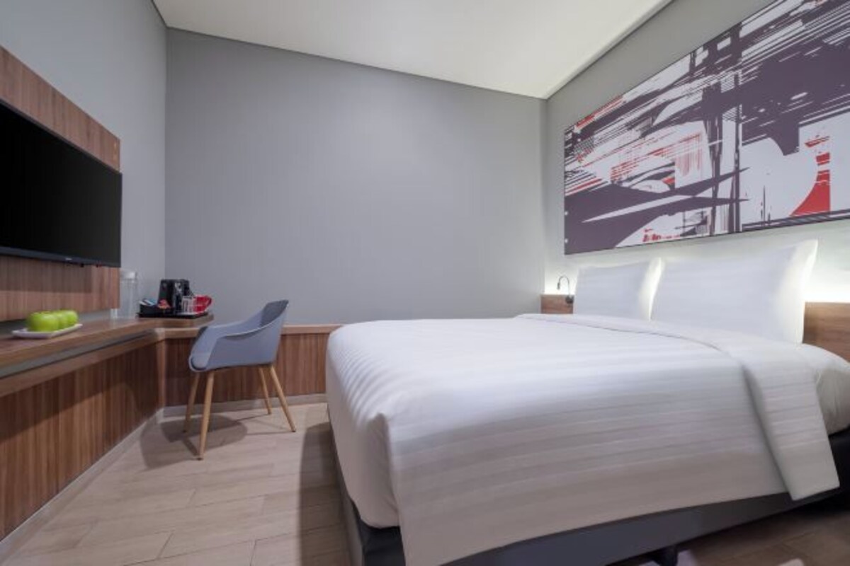 A modern hotel room with a large bed, a flat-screen TV mounted on the wall, a small desk with a chair, and a large abstract painting above the bed.