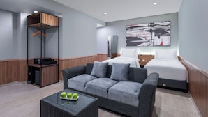 Habitación de hotel moderna con un sofá gris, una cama con ropa de cama blanca, un armario de madera y una obra de arte colgada en la pared.