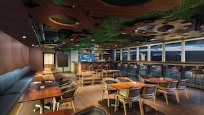 Un restaurante moderno con mesas y sillas de madera, una zona de bar y vistas a la ciudad a través de amplias ventanas.