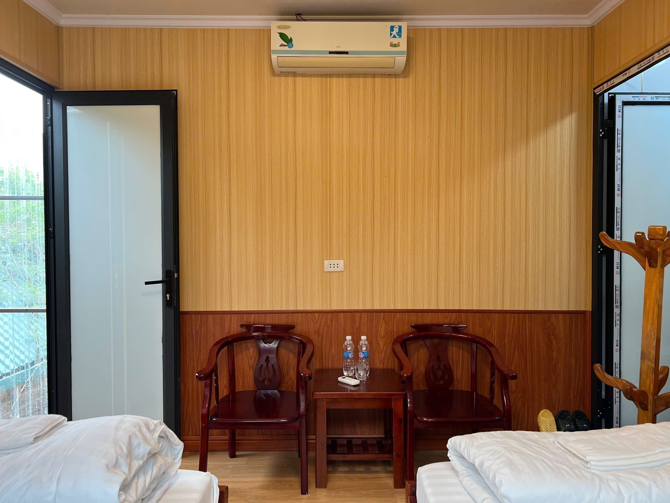 A hotel room with two beds, a wooden wall, an air conditioning unit, a small table with water bottles, and a wooden chair.