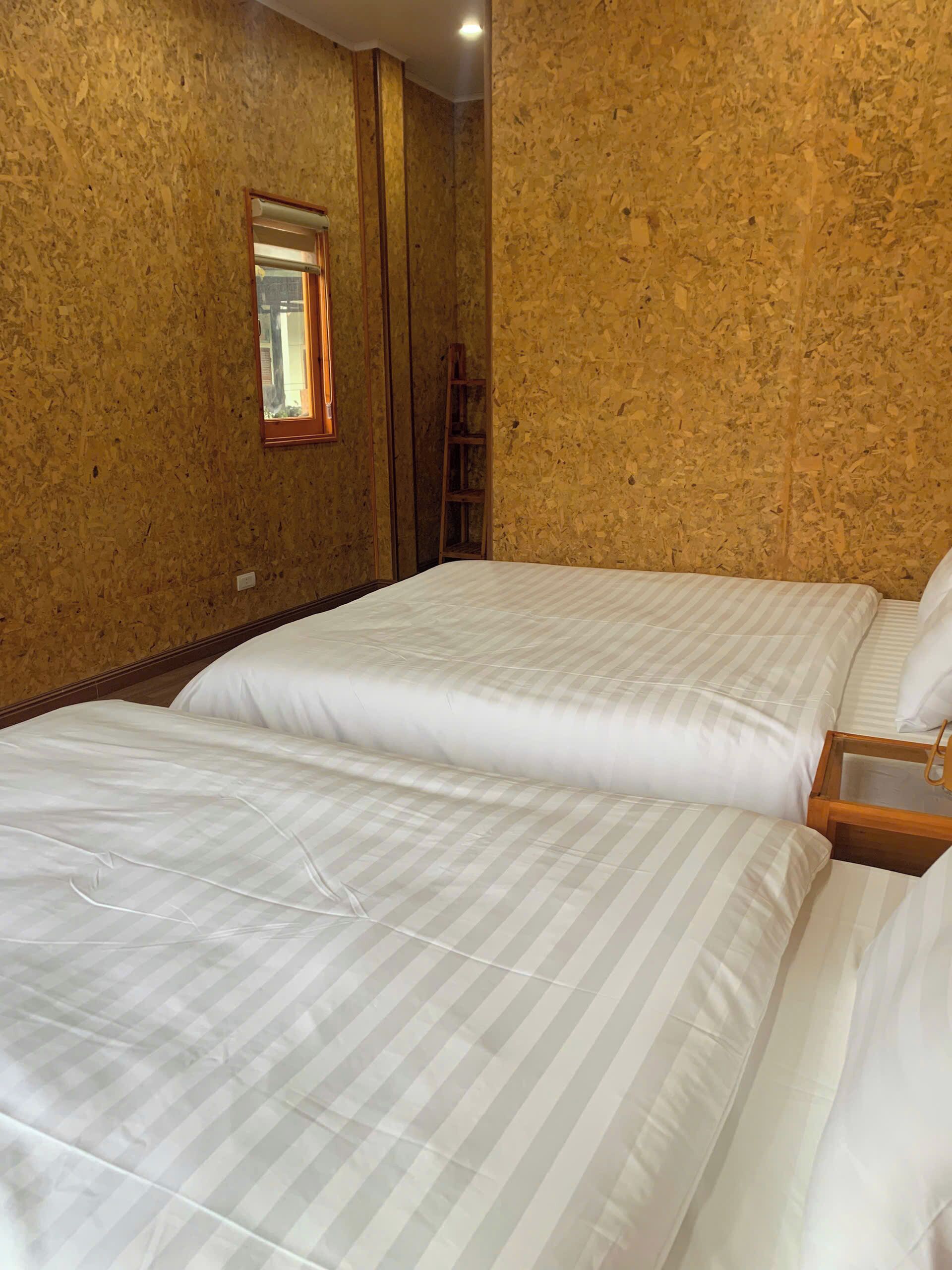 Ett hotellrum med två sängar, sänggavlar i trä, ett nattduksbord i trä, ett fönster med persienner och en trästege som står mot väggen.