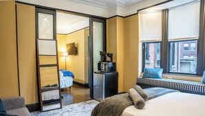 A hotel room with a bed, a seating area with a bench, a coffee machine, and a view of the city through large windows.