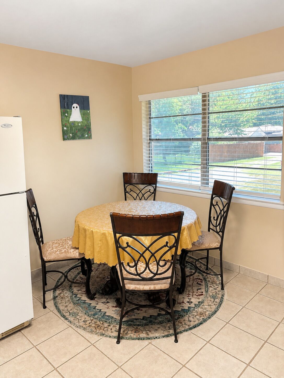 A dining area with a round table, four chairs, a refrigerator, a painting, and a window with blinds.