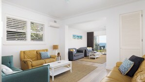 Un salon moderne avec un canapé jaune, un fauteuil bleu et un canapé gris. On y trouve une table basse blanche et une lampe. Les murs sont blancs et la pièce est dotée d’une grande fenêtre avec des stores.