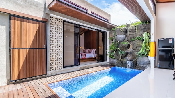A modern outdoor pool area with a wooden deck, a stone wall with a waterfall, and a sliding glass door leading to a bedroom. - Eden Villas Ubud (Ubud)