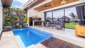 A modern outdoor living area with a swimming pool, wooden deck, and a stone wall with plants. - Eden Villas Ubud (Ubud)