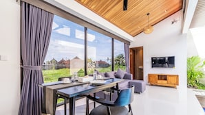 A modern living room with a dining area, large windows, a flat-screen TV, and a wooden ceiling. - Eden Villas Ubud (Ubud)