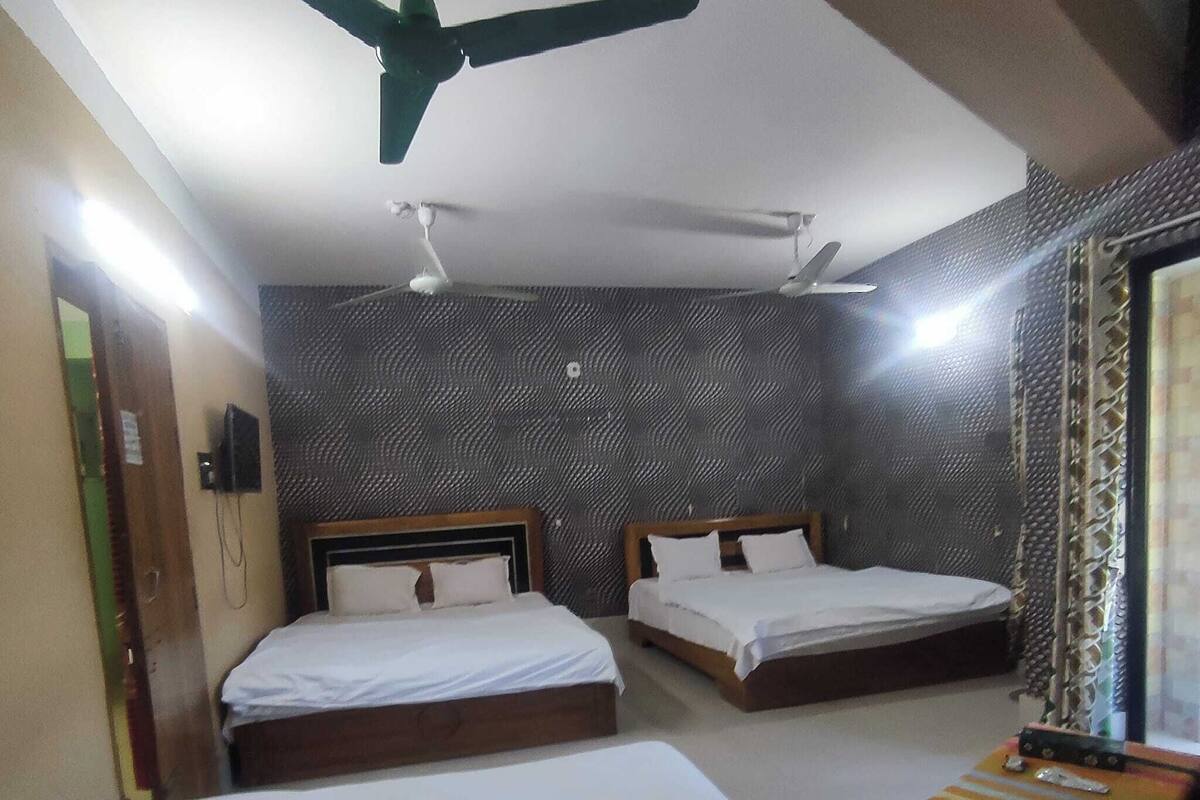 A hotel room with two single beds, a ceiling fan, a small table with a colorful cushion, and a patterned wall.