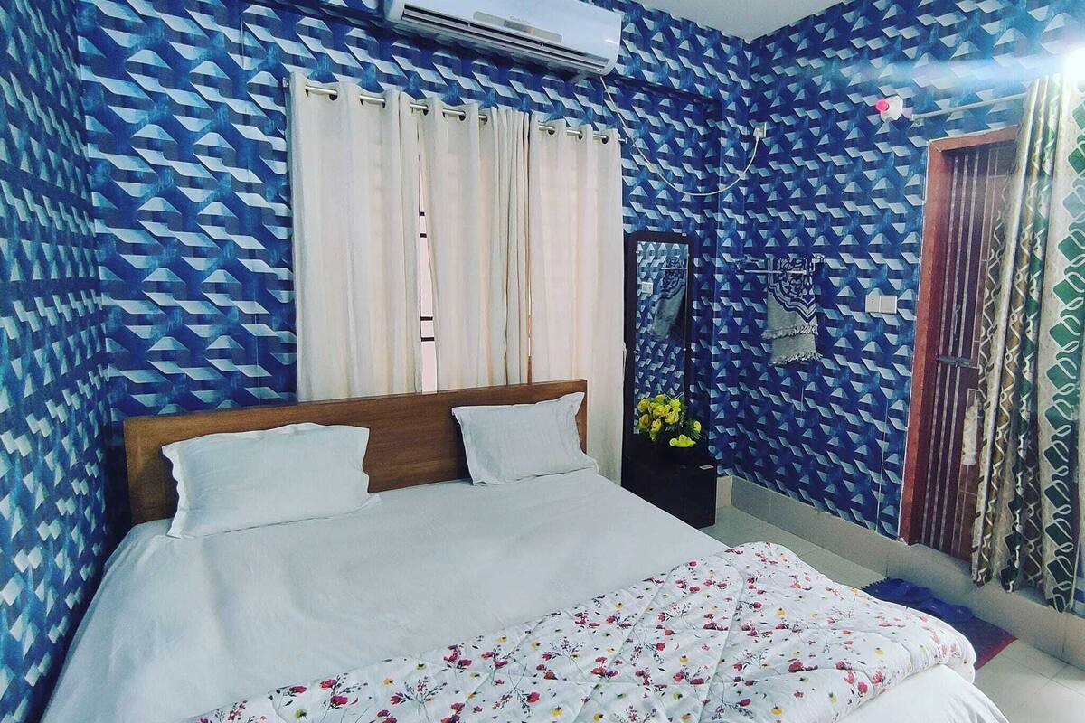 A bedroom with a bed, curtains, a nightstand, and a wall-mounted air conditioner.
